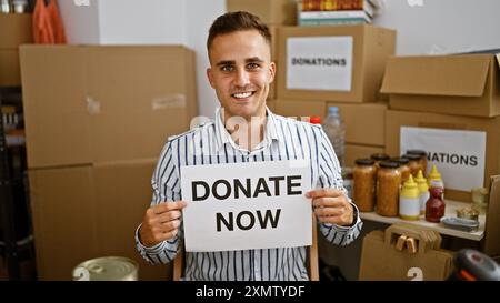 Un jeune homme souriant tenant une pancarte « donnez maintenant » dans un entrepôt rempli de boîtes de dons et de fournitures révèle une atmosphère caritative. Banque D'Images