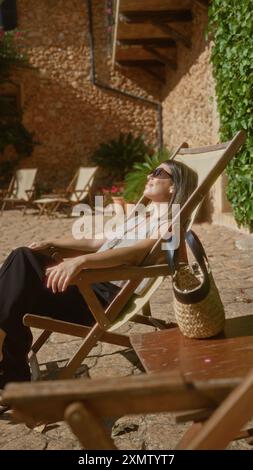 Femme se relaxant à l'extérieur par une journée ensoleillée à majorque en espagne, profitant d'un moment serein tout en étant assis sur une chaise longue dans une cour rustique Banque D'Images