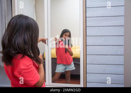 La fille de l'école primaire se pointe vers elle dans le miroir tout en chantant devant le miroir en utilisant un peigne comme microphone Banque D'Images