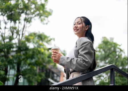 Une belle, attrayante femme d'affaires asiatique est debout à l'extérieur avec une tasse de café à emporter dans sa main, souriant et regardant loin avec un e rêveur éveillé Banque D'Images