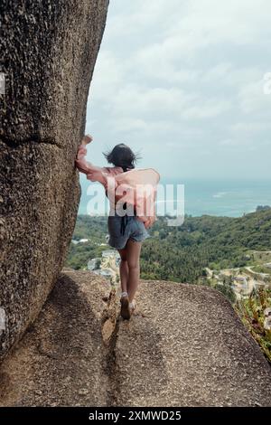 Une femme marche le long d'une étroite falaise, le dos à la caméra, avec l'océan bleu et la verdure tropicale derrière elle. Banque D'Images