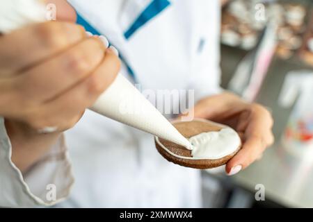 Gros plan : mains d'une femme chef pâtissière décorant des biscuits avec un glaçage blanc tout en pressant la crème hors d'un sac à passepoil. Photo de haute qualité Banque D'Images