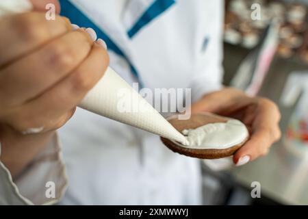 Gros plan : mains d'une femme chef pâtissière décorant des biscuits avec un glaçage blanc tout en pressant la crème hors d'un sac à passepoil. Photo de haute qualité Banque D'Images