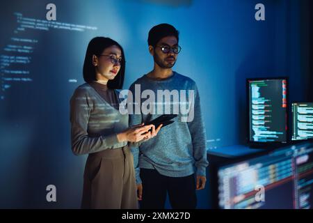 Asiatique deux développeurs de logiciels engagés dans une session de codage collaboratif, avec du code projeté sur le mur et plusieurs écrans d'ordinateur s'affichant Banque D'Images