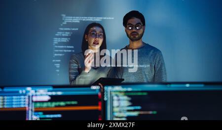 Asiatique deux développeurs de logiciels engagés dans une session de codage collaboratif, avec du code projeté sur le mur et plusieurs écrans d'ordinateur s'affichant Banque D'Images
