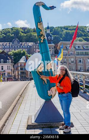 Dinant, Namur, Wallonie, Belgique. 5 juin 2024. Touriste féminine posant à côté d'une sculpture de saxophone peinte avec carte du monde, bâtiments sur brouillard brouillé b Banque D'Images