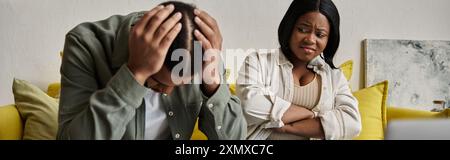 Couple afro-américain inquiet assis sur le canapé, homme contrarié, femme le regardant. Banque D'Images