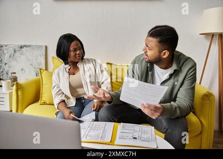 Un couple afro-américain aimant discute de leurs finances tout en étant assis sur un canapé jaune à la maison. Banque D'Images