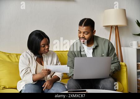 Un couple afro-américain est assis sur un canapé jaune, examinant des documents et utilisant un ordinateur portable. Banque D'Images