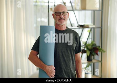 Portrait d'un homme caucasien âgé souriant tenant un tapis de yoga tout en se tenant debout dans la chambre avec des plantes et des étagères, portant des lunettes et des vêtements décontractés Banque D'Images