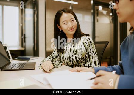 Jeune manager asiatique assis à son bureau et regardant son collègue masculin tout en travaillant avec des documents ensemble Banque D'Images