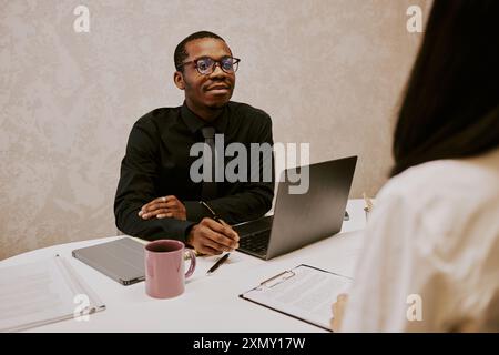 Photo au-dessus de l'épaule d'un jeune responsable RH afro-américain assis à son bureau parlant à une femme méconnaissable Banque D'Images