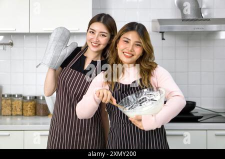 Deux femmes dans des tabliers souriant dans la cuisine, l'une tenant un gant de four, l'autre mélangeant la pâte dans un bol. Banque D'Images