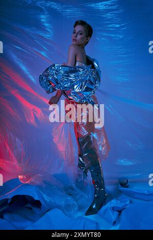 Une femme dans une veste argentée et des bottes noires pose au milieu d'une mer tourbillonnante de plastique bleu et rouge. Banque D'Images