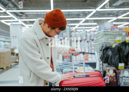 Homme en manteau confortable et bonnet examine la valise rouge au magasin, se préparant pour des voyages ou des voyages de vacances. Masculin choisit la valise pour les vacances au supermarché. Banque D'Images