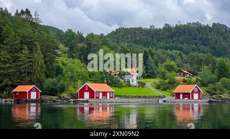 Les cabanes rouges vibrantes se reflètent sur les eaux calmes sous un ciel nuageux, entourées de forêts denses et de collines en Norvège. Banque D'Images