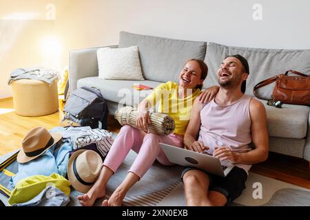 Jeune couple est assis sur le sol à la maison et s'amuse tout en réservant des vols et des hôtels. Préparez-vous pour les vacances d'été. Banque D'Images