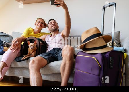 Le jeune couple est assis sur le canapé avec leur chien Toy Terrier et prend des photos avant ou après le voyage. Banque D'Images