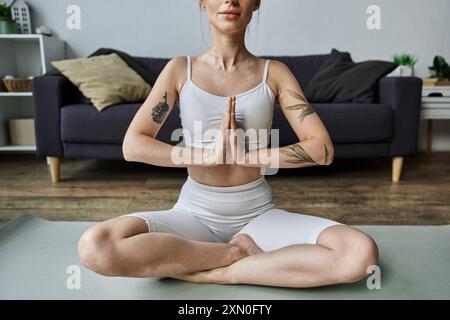 Une jeune femme est assise dans une pose de yoga méditative sur un tapis, portant des vêtements de sport blancs dans un cadre d'appartement moderne. Banque D'Images