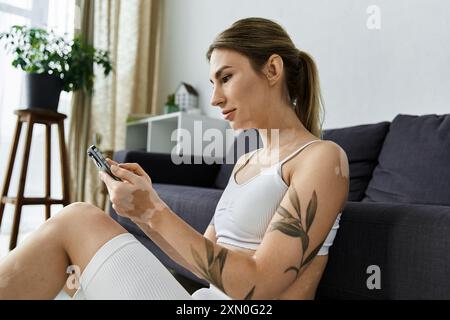 Une jeune femme en vêtements d'action est assise sur le sol d'un appartement moderne, à l'aide de son téléphone. Banque D'Images