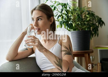 Une jeune femme atteinte de vitiligo, vêtue de vêtements actifs, est assise dans un appartement moderne et boit de l'eau après avoir pratiqué le yoga. Banque D'Images