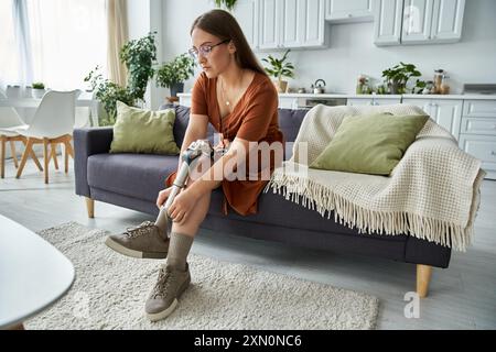 Une femme est assise sur un canapé dans un salon, ajustant sa jambe prothétique. Banque D'Images