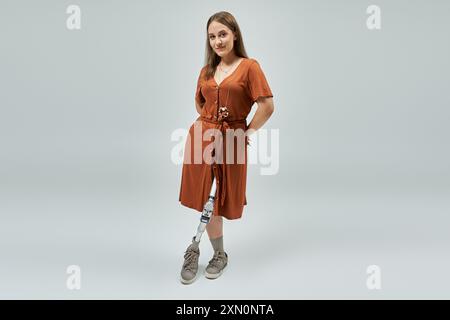 Une femme avec une jambe prothétique se tient confiante dans une robe de couleur rouille sur un fond Uni. Banque D'Images