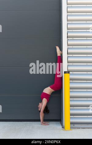 Femme sportive faisant un stand contre un mur industriel, portant des vêtements de sport rouges Banque D'Images