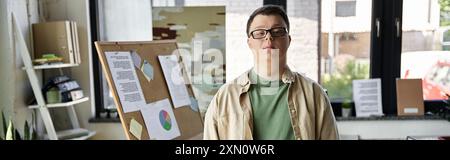Un jeune homme atteint du syndrome de Down se tient confiant devant un tableau d'affichage dans un bureau. Banque D'Images
