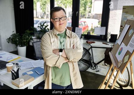 Un jeune homme atteint du syndrome de Down se tient confiant avec les bras croisés dans un espace de bureau lumineux. Banque D'Images