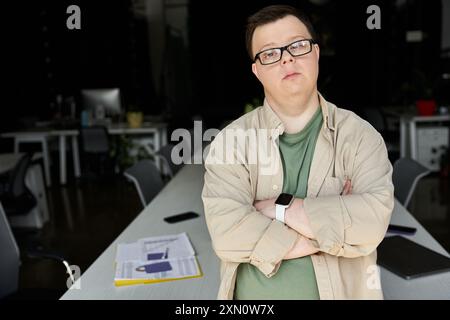 Un homme atteint du syndrome de Down se tient confiant dans un bureau avec les bras croisés. Banque D'Images