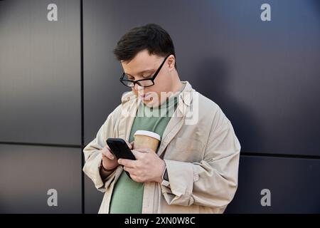 Un homme atteint du syndrome de Down se tient près d'un mur, regardant son téléphone tout en tenant une tasse de café. Banque D'Images