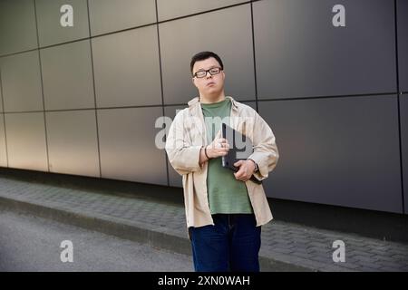 Un jeune homme atteint du syndrome de Down se tient confiant, tenant un ordinateur portable devant un bâtiment gris. Banque D'Images