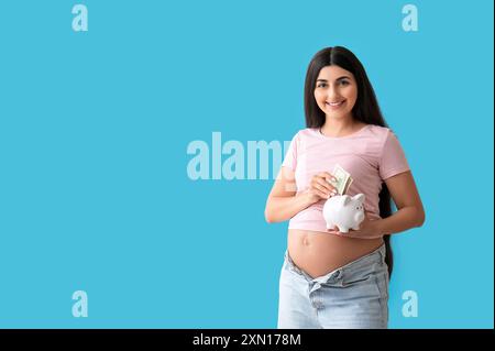 Jeune femme enceinte mettant de l'argent dans la tirelire sur fond bleu. Concept de prestations maternelles Banque D'Images