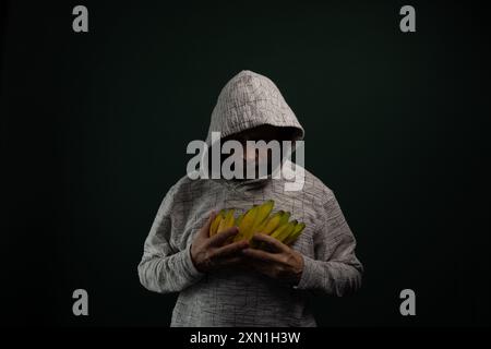 Homme mystérieux portant un sweat à capuche tenant un bouquet de bananes jaunes. Isolé sur fond sombre. Banque D'Images