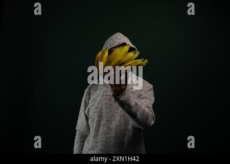 Homme mystérieux portant un sweat à capuche tenant un bouquet de bananes jaunes devant son visage. Isolé sur fond sombre. Banque D'Images