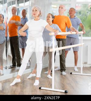 Femme âgée active joyeuse formant diverses poses de ballet pendant des cours de groupe avec des danseurs de ballet à la barre dans le studio de chorégraphie Banque D'Images