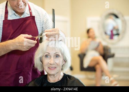 Le maître expérimenté de l'homme senior fait effectue la coupe de cheveux pour la femme mature avec des ciseaux Banque D'Images