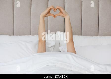 Bonjour. Femme faisant un geste de coeur avec les mains dans le lit, gros plan Banque D'Images