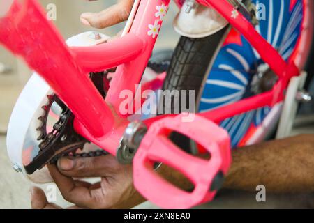 Gros plan des mains assemblant ou fixant un vélo d'enfant rose vif, parfait pour les thèmes du bricolage, de l'enfance et de la famille. Banque D'Images