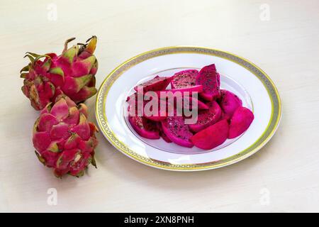 Délicieuses tranches de fruits de dragon sur une assiette blanche sur fond en bois. Deux fruits de dragon entiers placés à côté de l'assiette. Banque D'Images