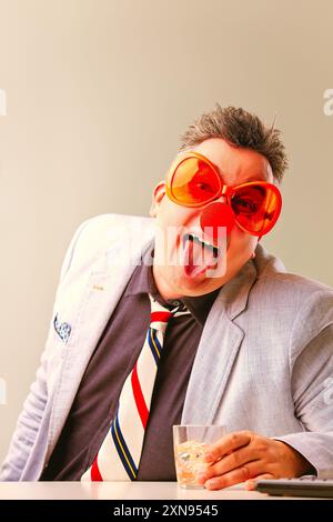 Homme d'affaires dans des verres surdimensionnés et un nez de clown sort de sa langue, tenant un verre d'eau, créant une atmosphère ludique et joyeuse Banque D'Images