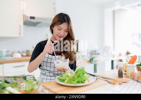 Jeune femme asiatique utilise une cuillère pour ajouter de la mayonnaise dans la salade sur l'assiette tout en préparant la salade de légumes frais dans un bol et en cuisinant des aliments sains pour le petit déjeuner Banque D'Images