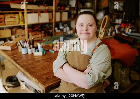 Jeune travailleuse d'atelier avec le syndrome de Down regardant la caméra et debout avec les bras croisés dans un vieil atelier entièrement équipé d'étagères et de matériel de couture Banque D'Images
