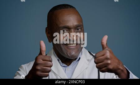Portrait homme afro-américain souriant médecin en manteau blanc montrant les pouces vers le haut dans un fond bleu studio regardant l'approbation du geste de la caméra Banque D'Images