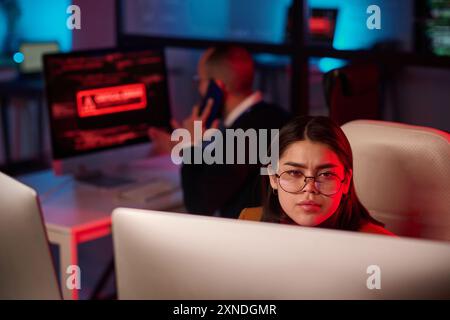Portrait de jeune femme portant des lunettes et regardant l'écran d'ordinateur dans le département de cybersécurité avec l'espace de copie de lumière d'urgence rouge Banque D'Images