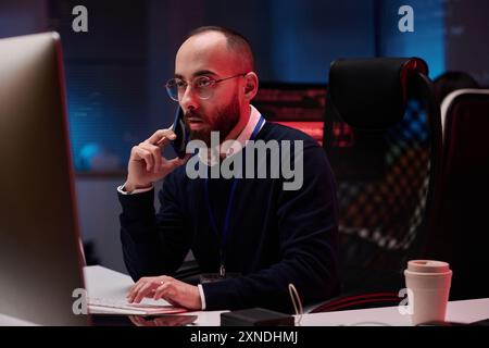 Portrait d'un homme adulte à l'aide d'un ordinateur et parlant au téléphone tout en travaillant sur l'urgence dans l'espace de copie du département de cybersécurité Banque D'Images