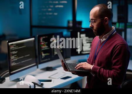 Portrait de vue latérale de l'homme adulte barbu comme programmeur informatique tenant un ordinateur portable ouvert dans LE bureau INFORMATIQUE et de cybersécurité avec l'espace de copie des lumières bleues Banque D'Images