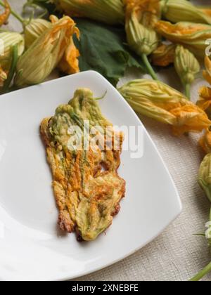 Vertical directement au-dessus gros plan de fleurs de courgettes frites dans la pâte sur une assiette blanche, entouré de fleurs de courgettes fraîches sur une nappe blanche. Banque D'Images