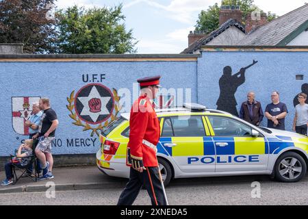 Ballyclare, Irlande du Nord - 27 août 2022 : Bandsman passant devant la murale paramilitaire de l'UFF et la voiture de police du PSNI, samedi dernier, Royal Black institution. Banque D'Images